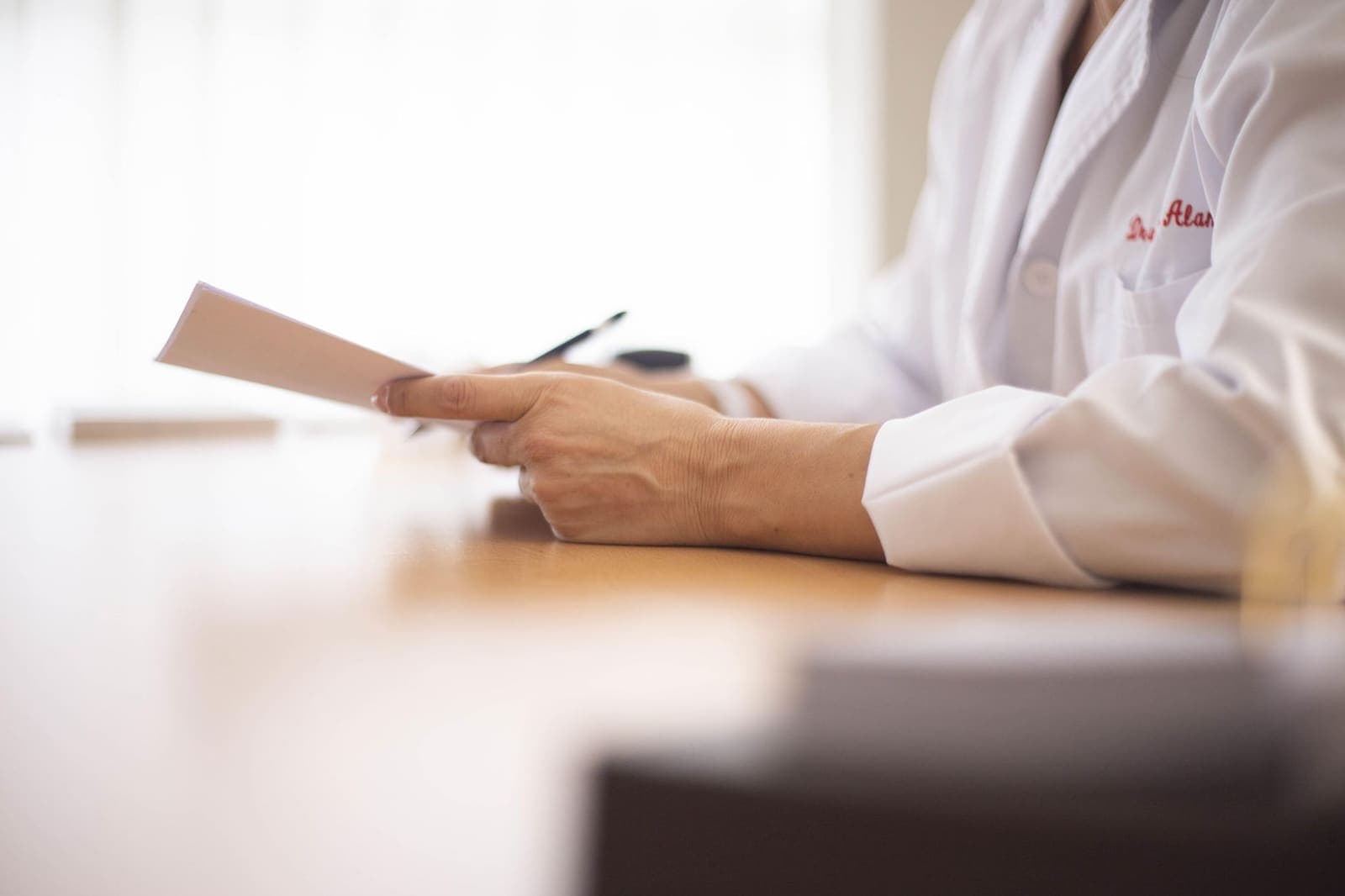 Close-up of a physician's hands in a white coat holding a sheet of paper and a pen at a wooden desk