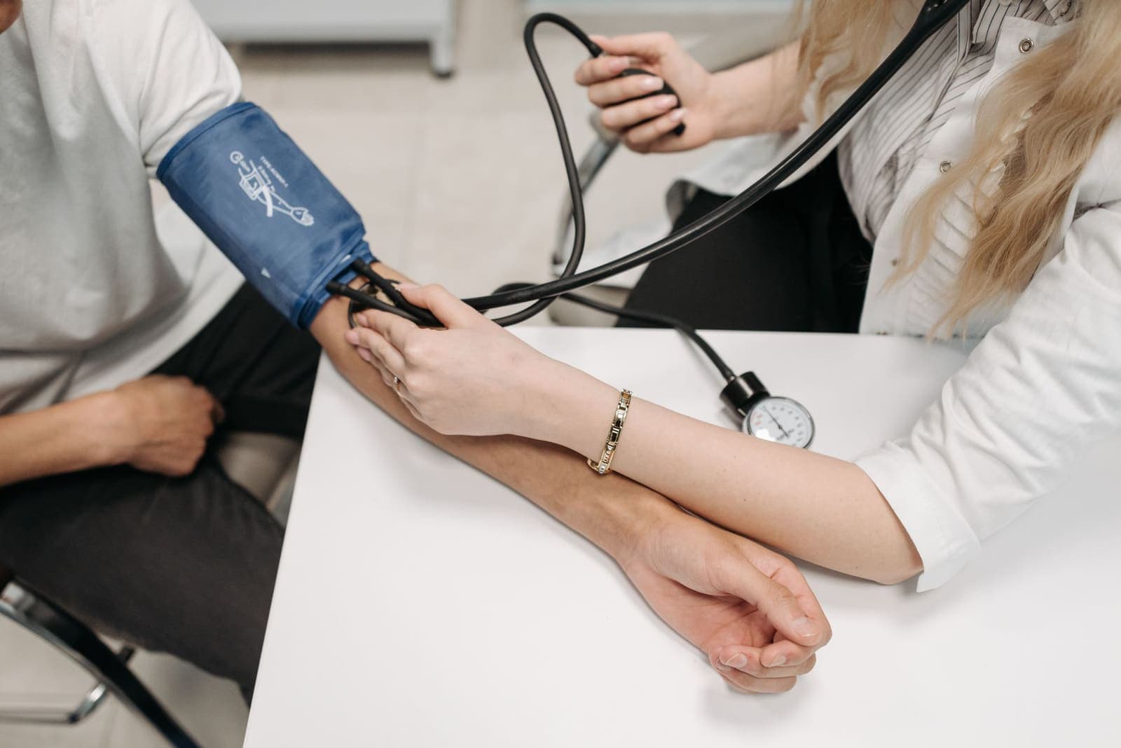 A clinician in a white coat taking a patient's blood pressure with a manual cuff and gauge on a white desk