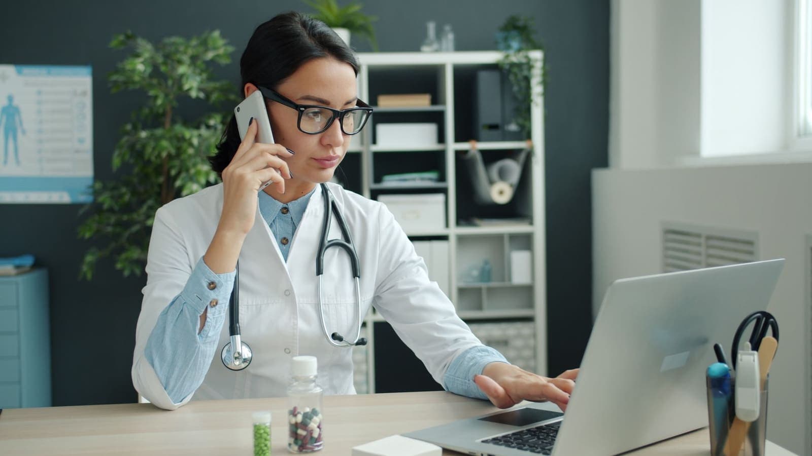 A doctor on the phone at a desk while working on a laptop