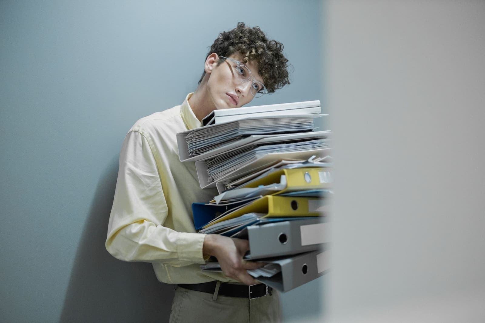 A young man in glasses leans against a wall holding a tall stack of overfilled binders and folders