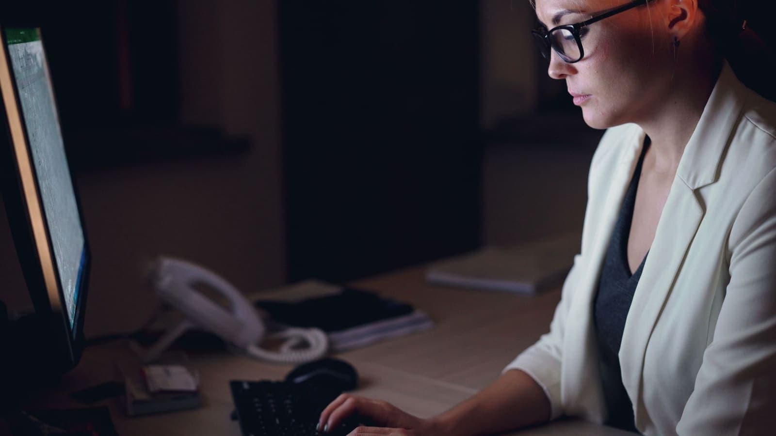 A woman in glasses and a white jacket types at a computer in a dark office after hours, a desk phone beside the keyboard