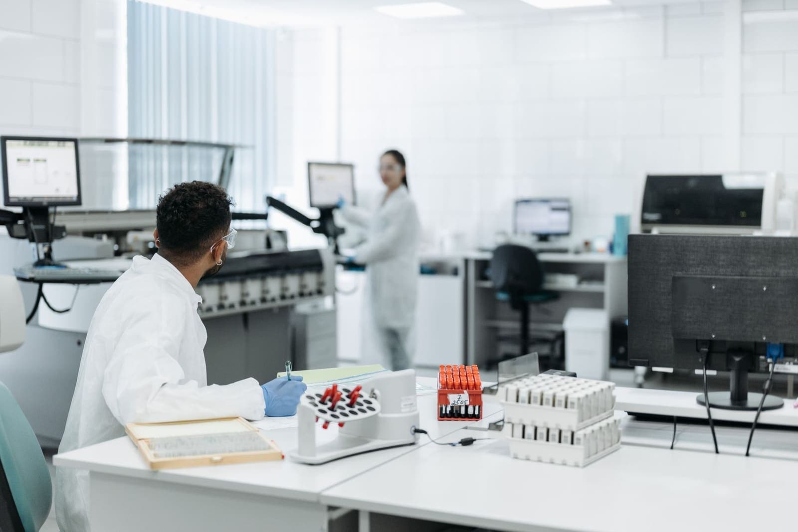 A laboratory technician in a white gown and gloves writes on a form at a bench beside racks of sample tubes