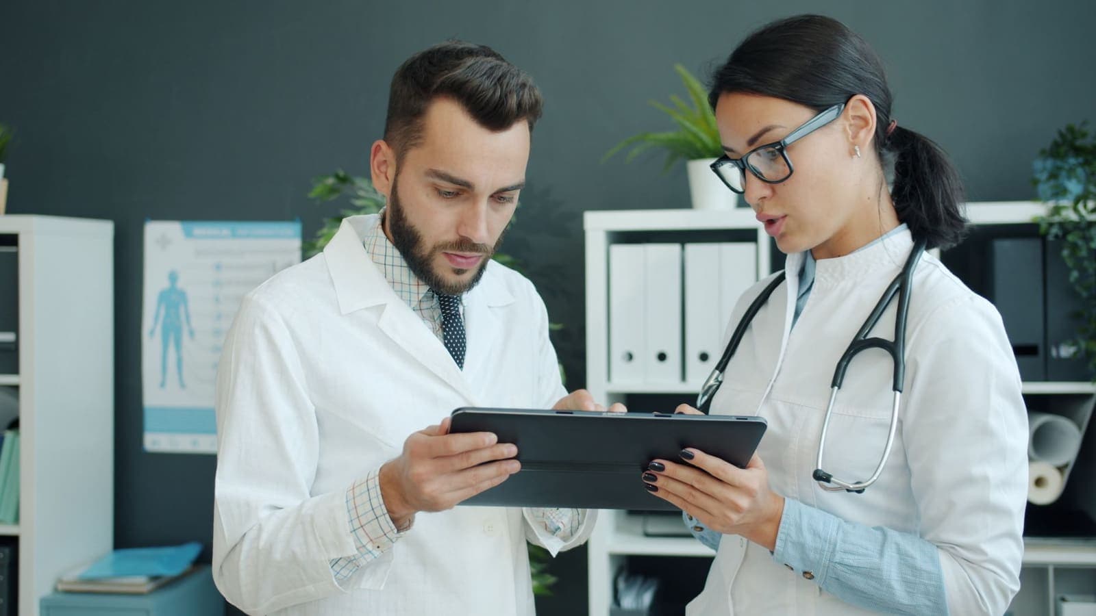 Two clinicians in white coats review a record together on a tablet in a clinic office