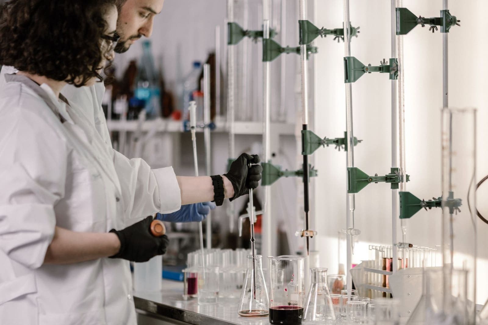Two researchers in lab coats and gloves work at a bench of flasks and burettes in a laboratory