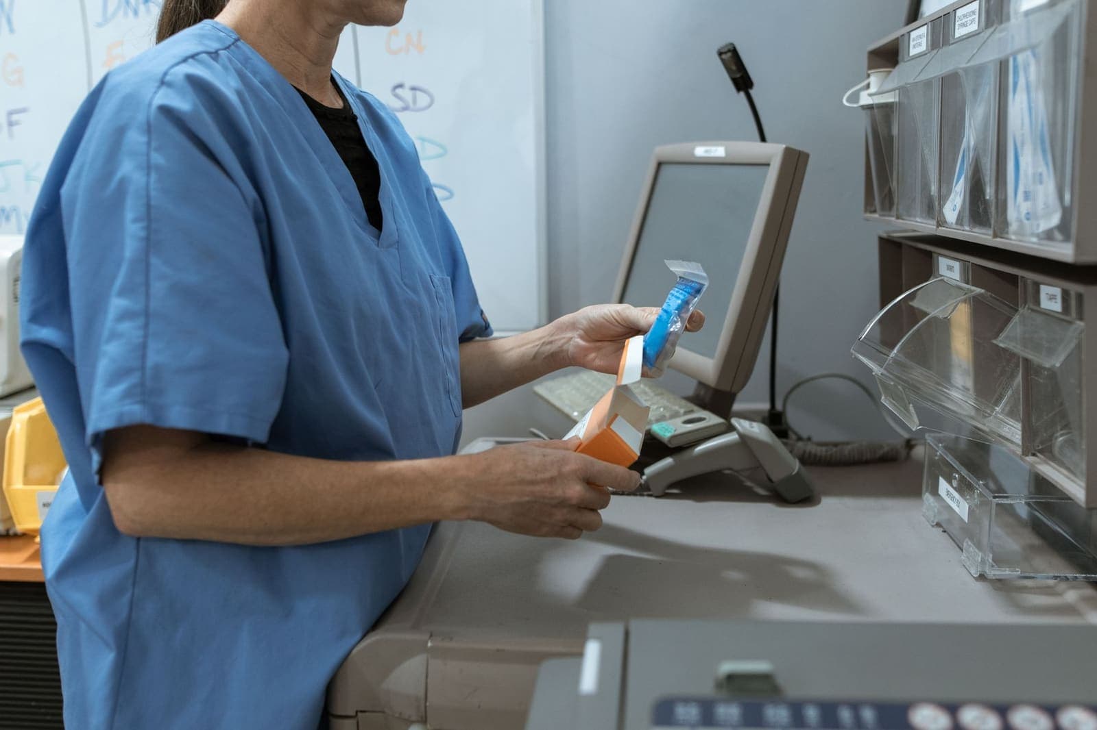 A healthcare worker in blue scrubs opens a medication package beside labelled supply bins and a computer