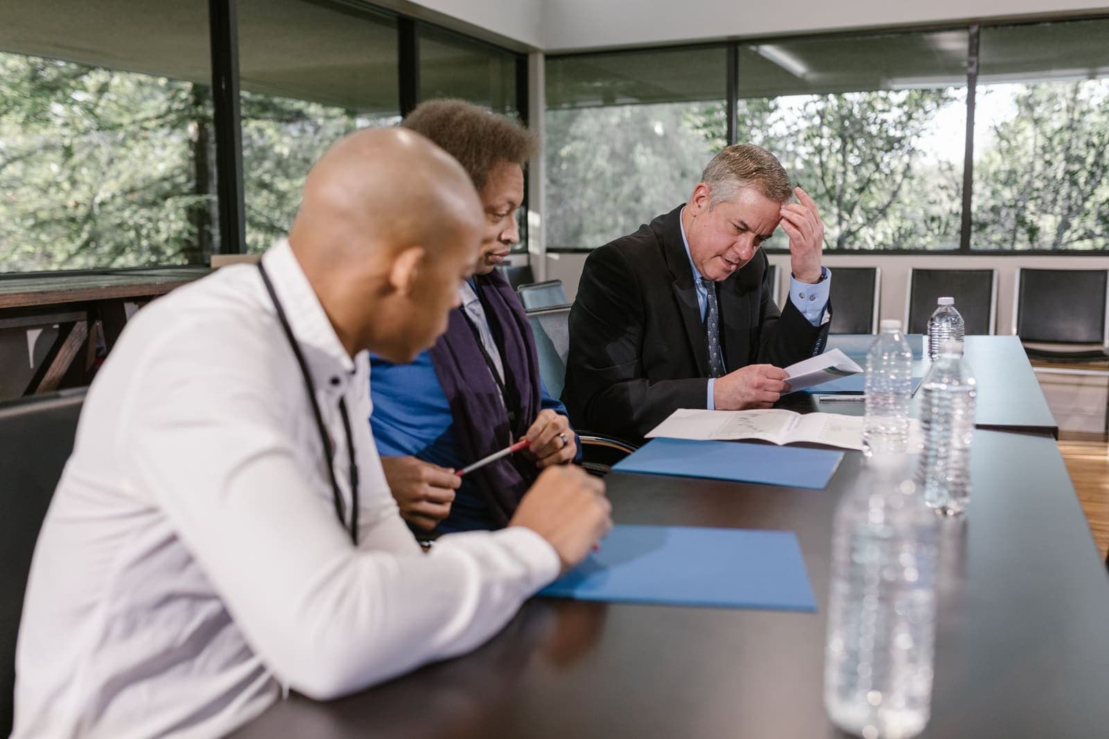 Three people sit at a conference table reviewing printed documents and blue folders beside a window