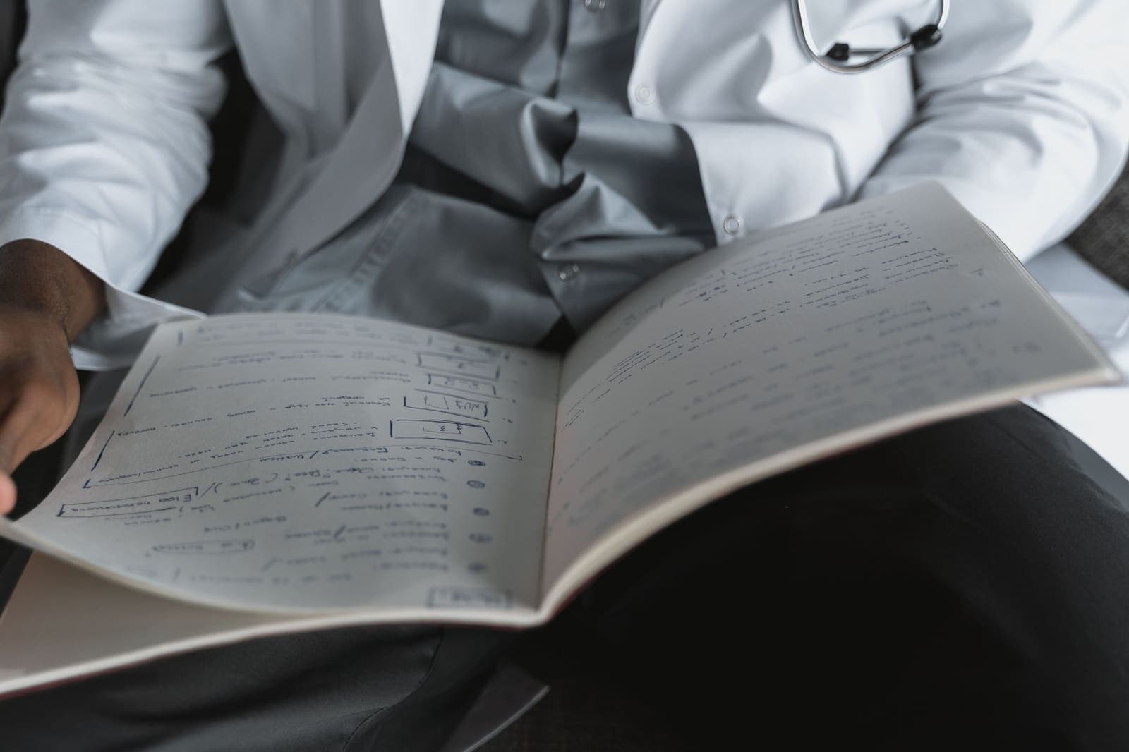A clinician in a white coat with a stethoscope reads handwritten notes in an open notebook