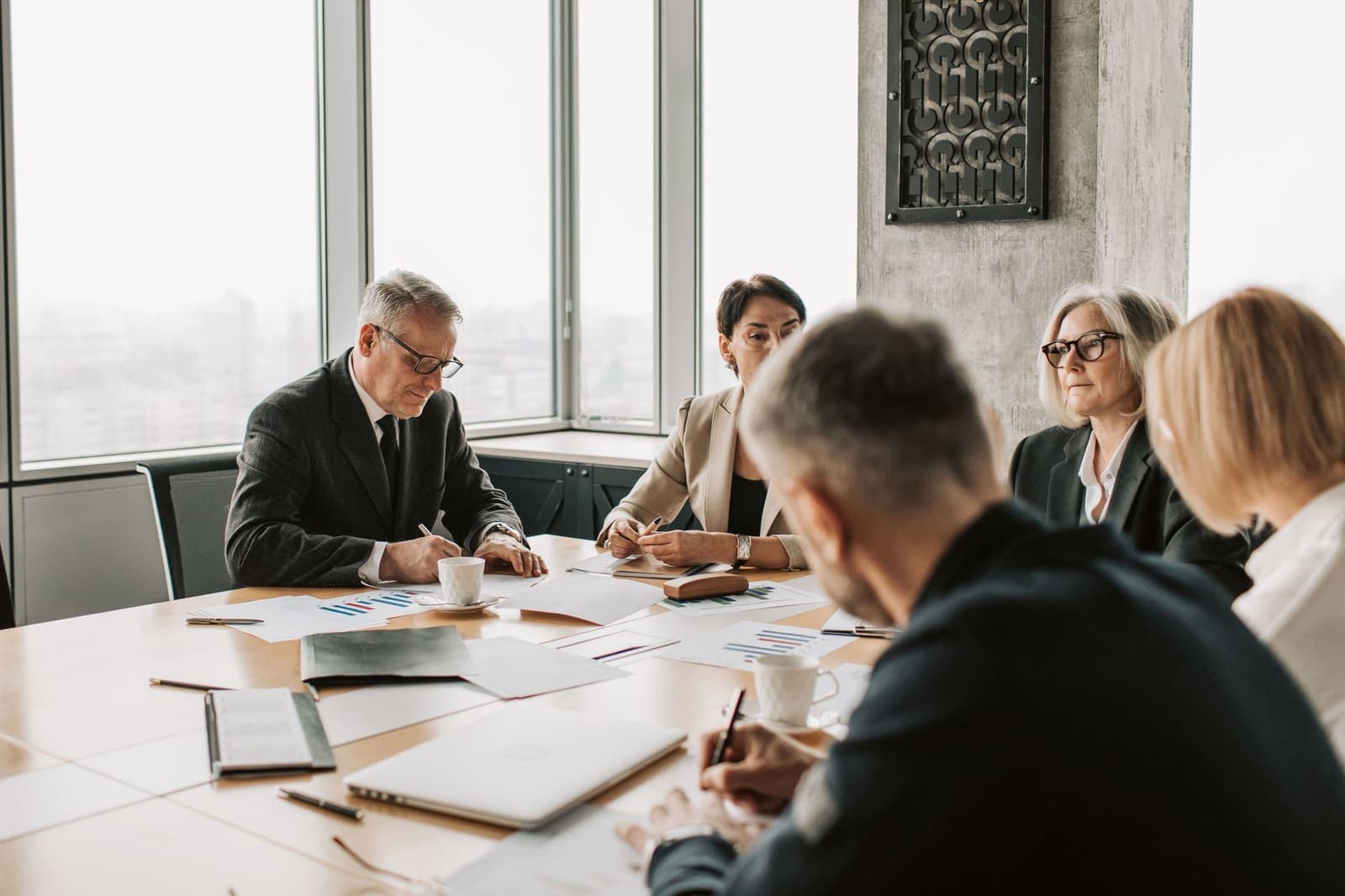 A committee of five people around a conference table with papers, charts and coffee cups in a high-rise meeting room