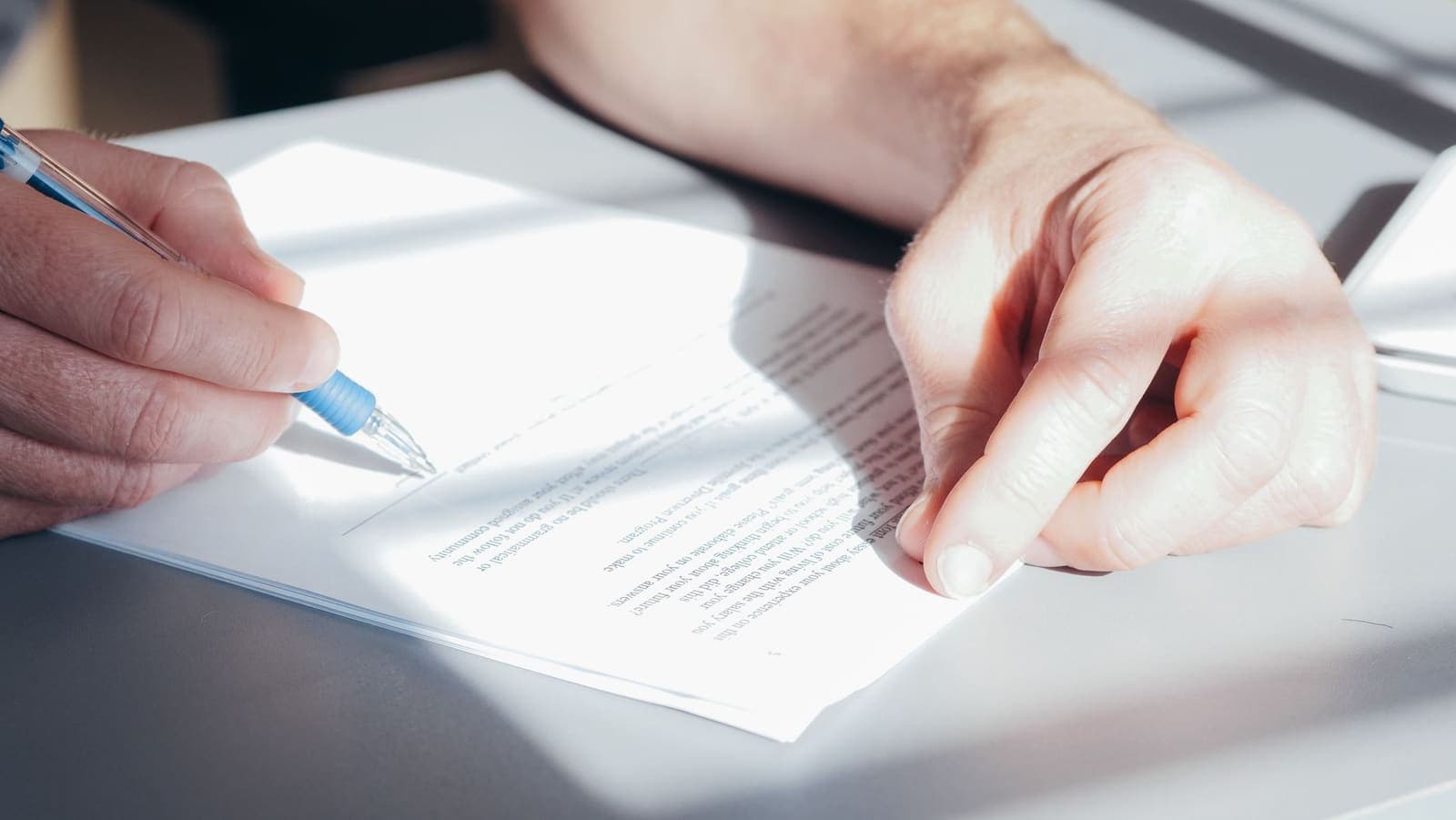 A hand signing a printed document with a blue ballpoint pen on a sunlit desk