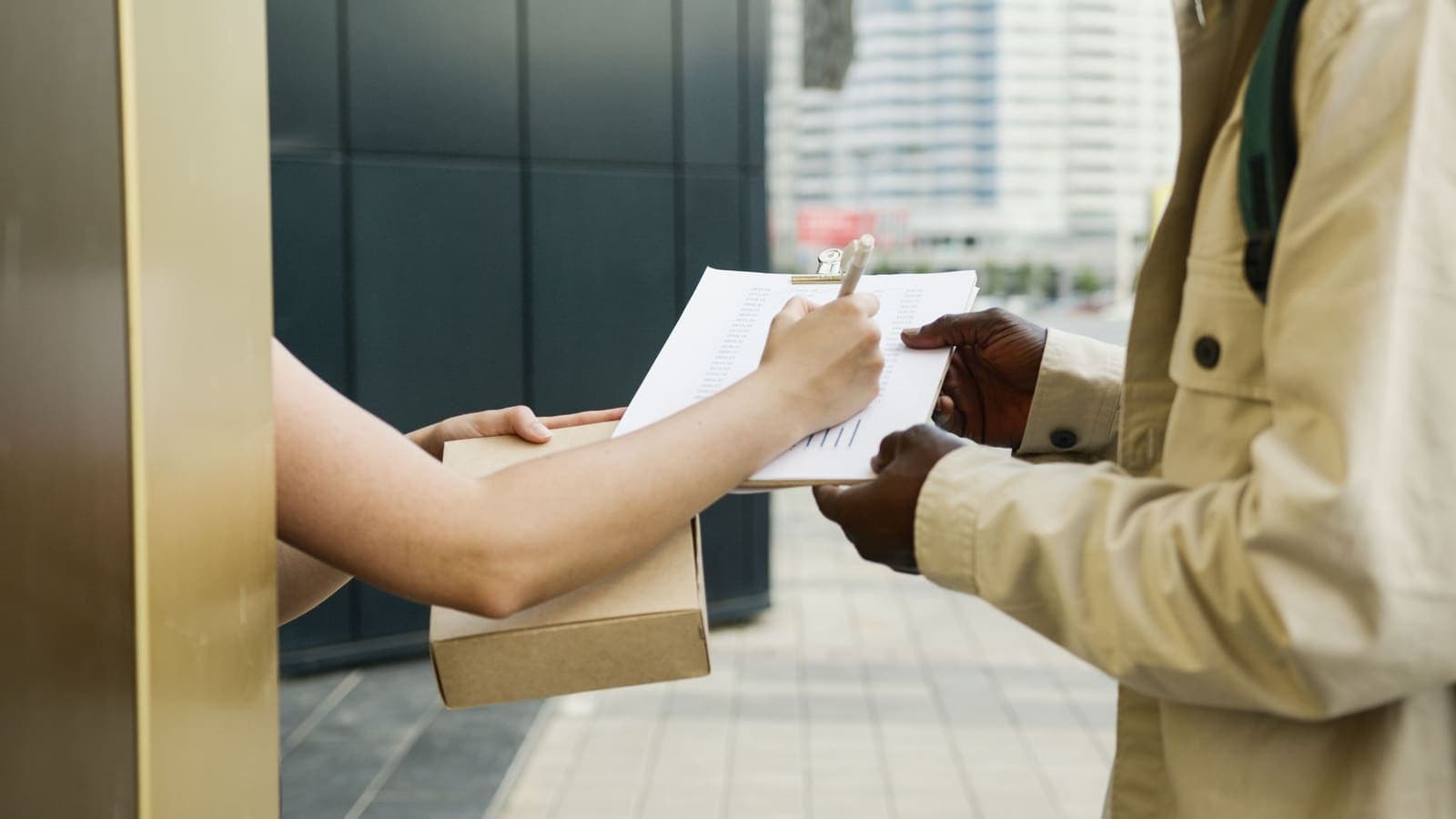 A person signs a courier's clipboard while holding a small cardboard parcel at a building entrance