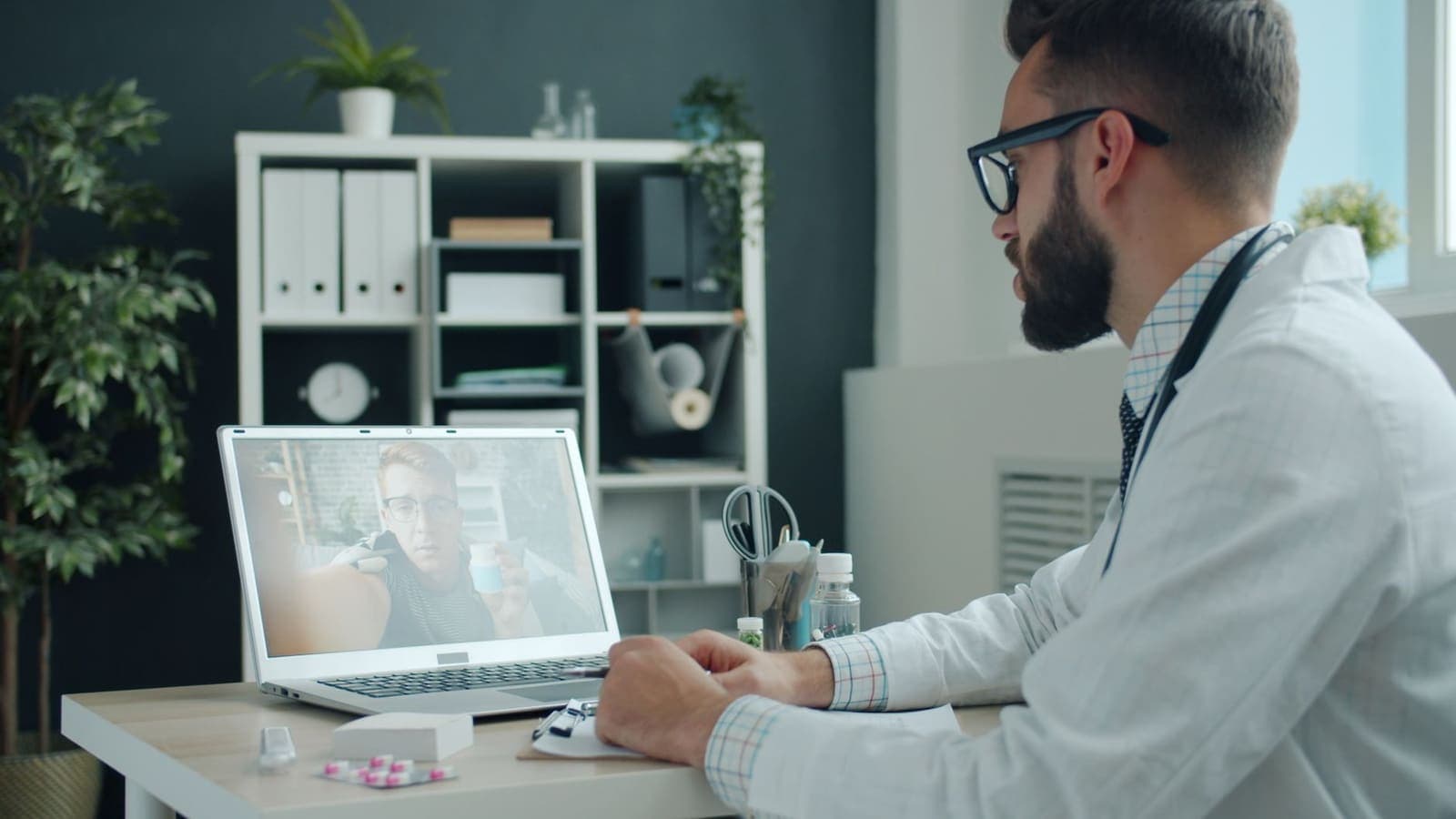 A clinician in a white coat takes notes during a video call with a patient shown on a laptop screen