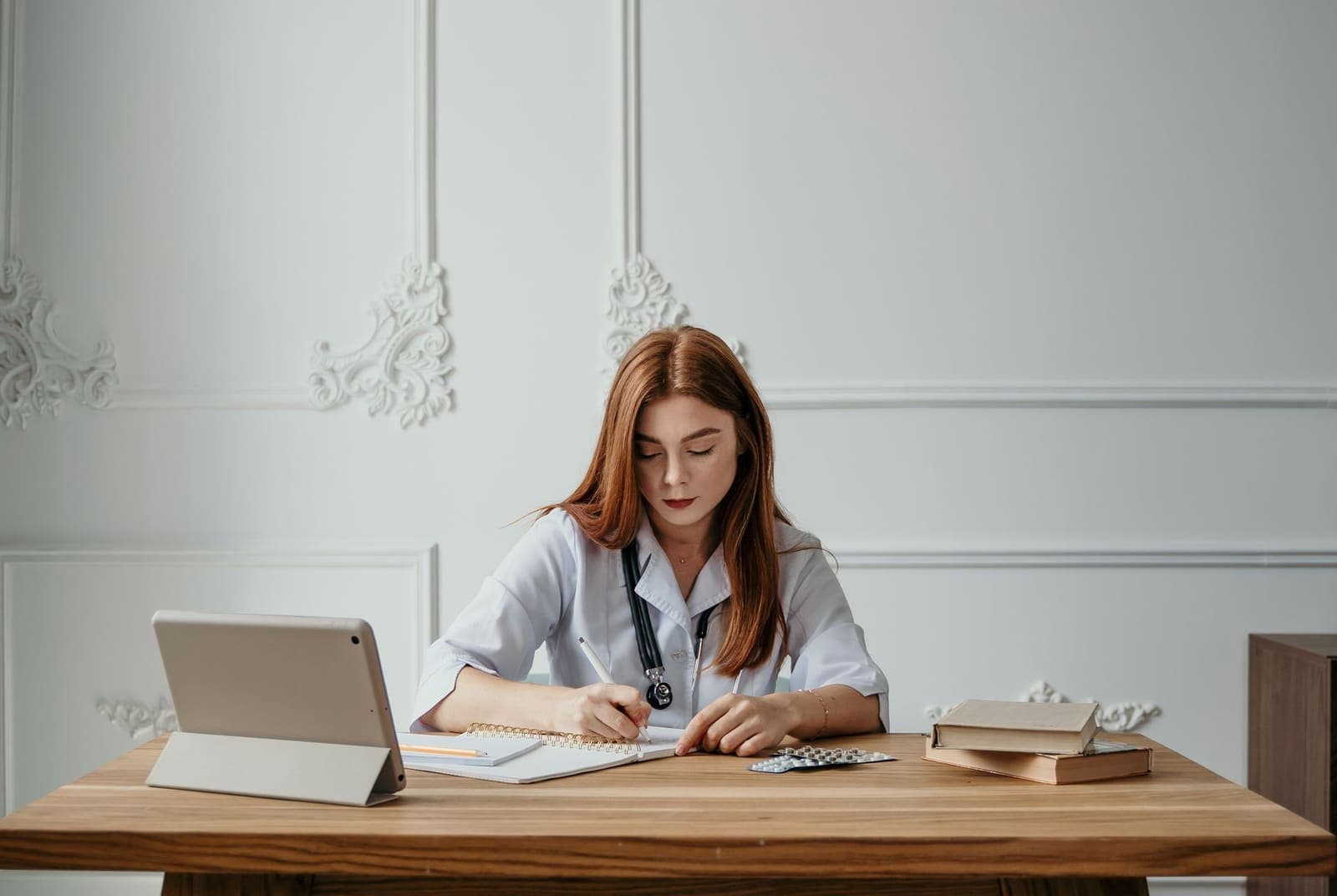 A clinician in a white coat writes in a notebook at a wooden desk with a tablet, books and a blister pack beside her