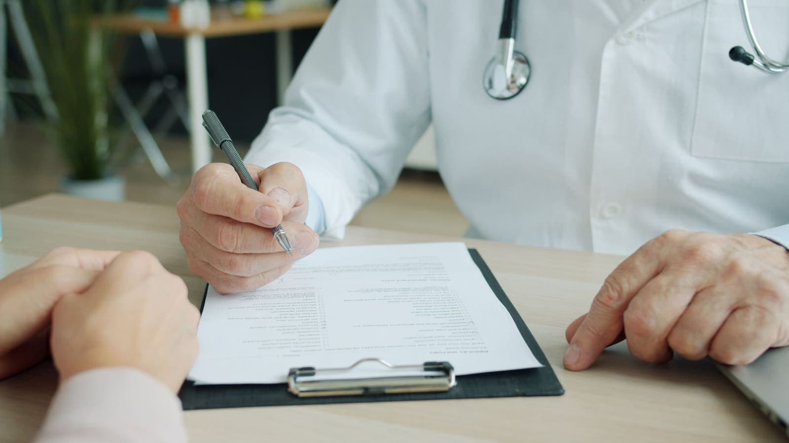 A physician in a white coat with a stethoscope fills in a form on a clipboard across a desk from a patient