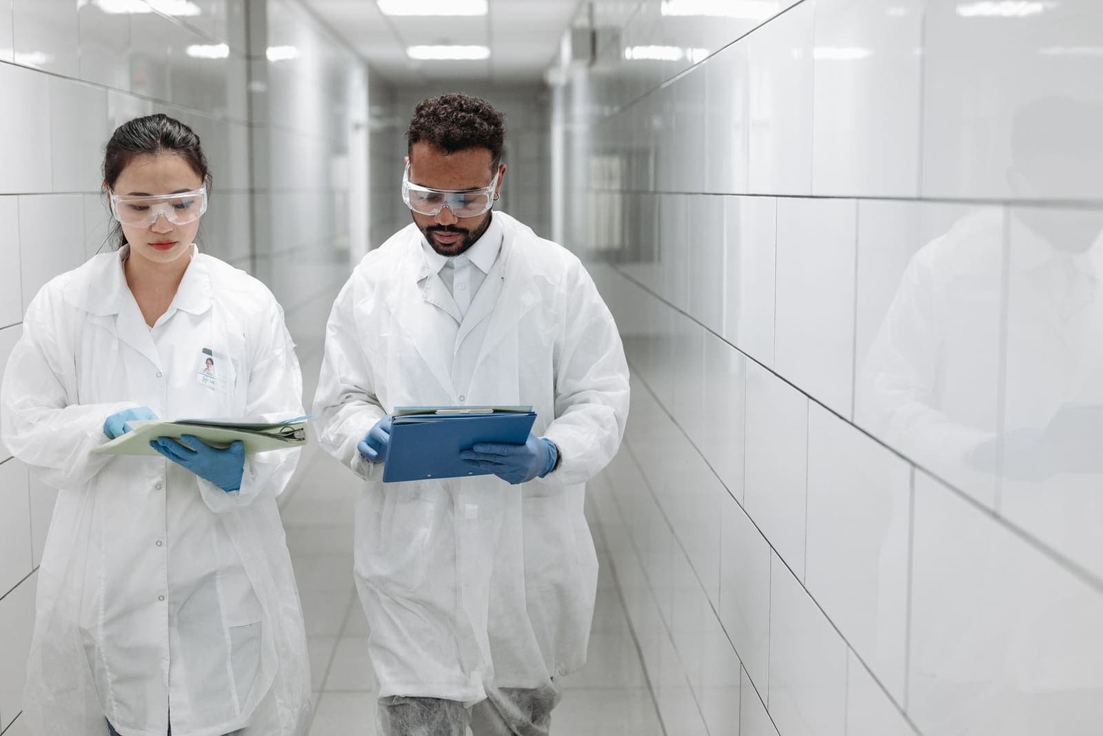 Two researchers in lab coats, safety glasses and blue gloves read folders as they walk down a white tiled corridor
