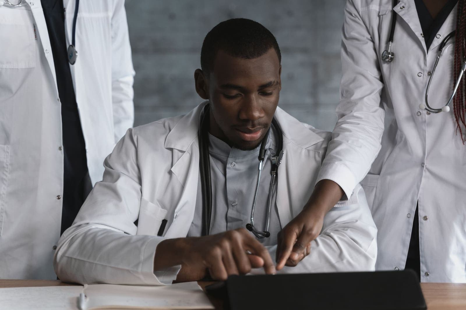 A physician at a desk works on a tablet while two colleagues in white coats stand beside him and one points at the screen