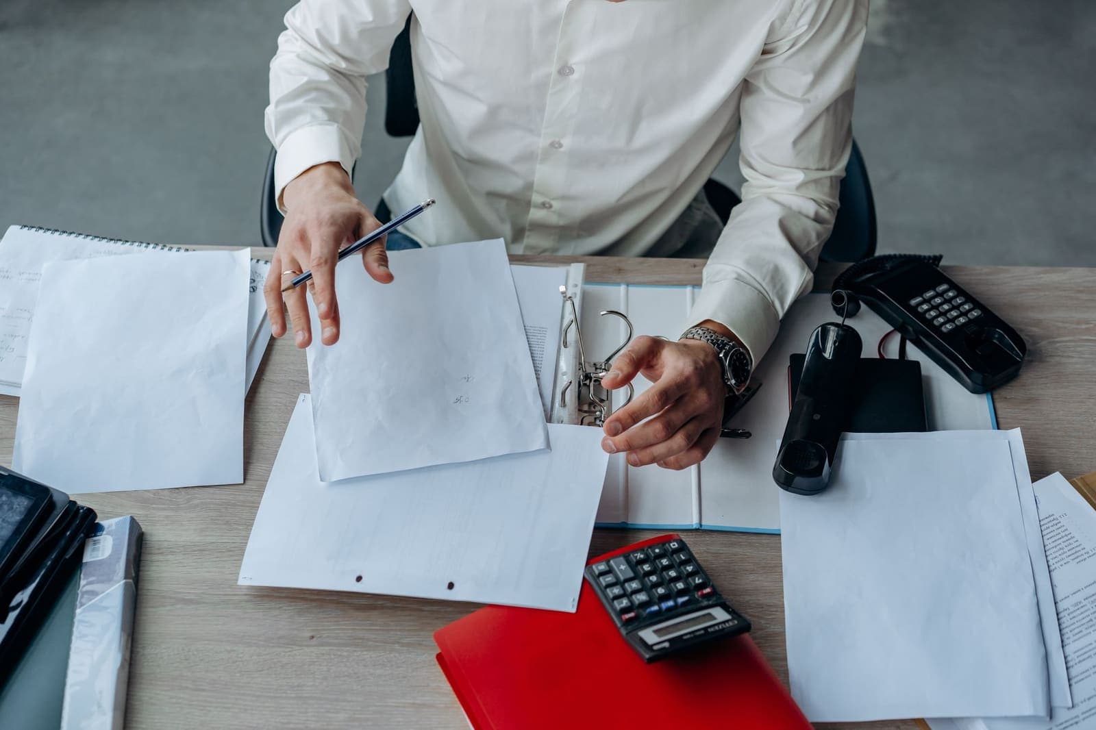 A man at a desk sorting loose pages into a ring binder beside a desk phone and calculator