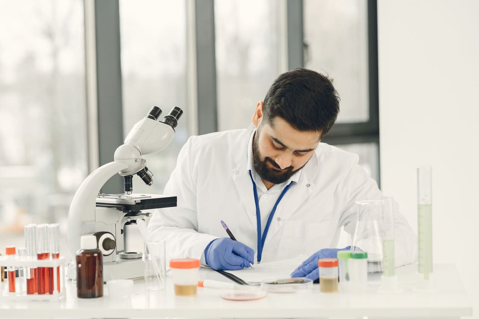 A researcher in a white coat and blue gloves writing notes at a lab bench beside a microscope and sample tubes
