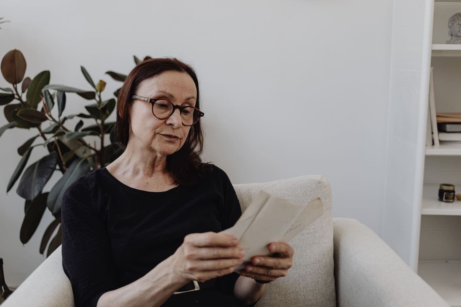 An older woman in glasses sitting in an armchair at home reading a folded letter