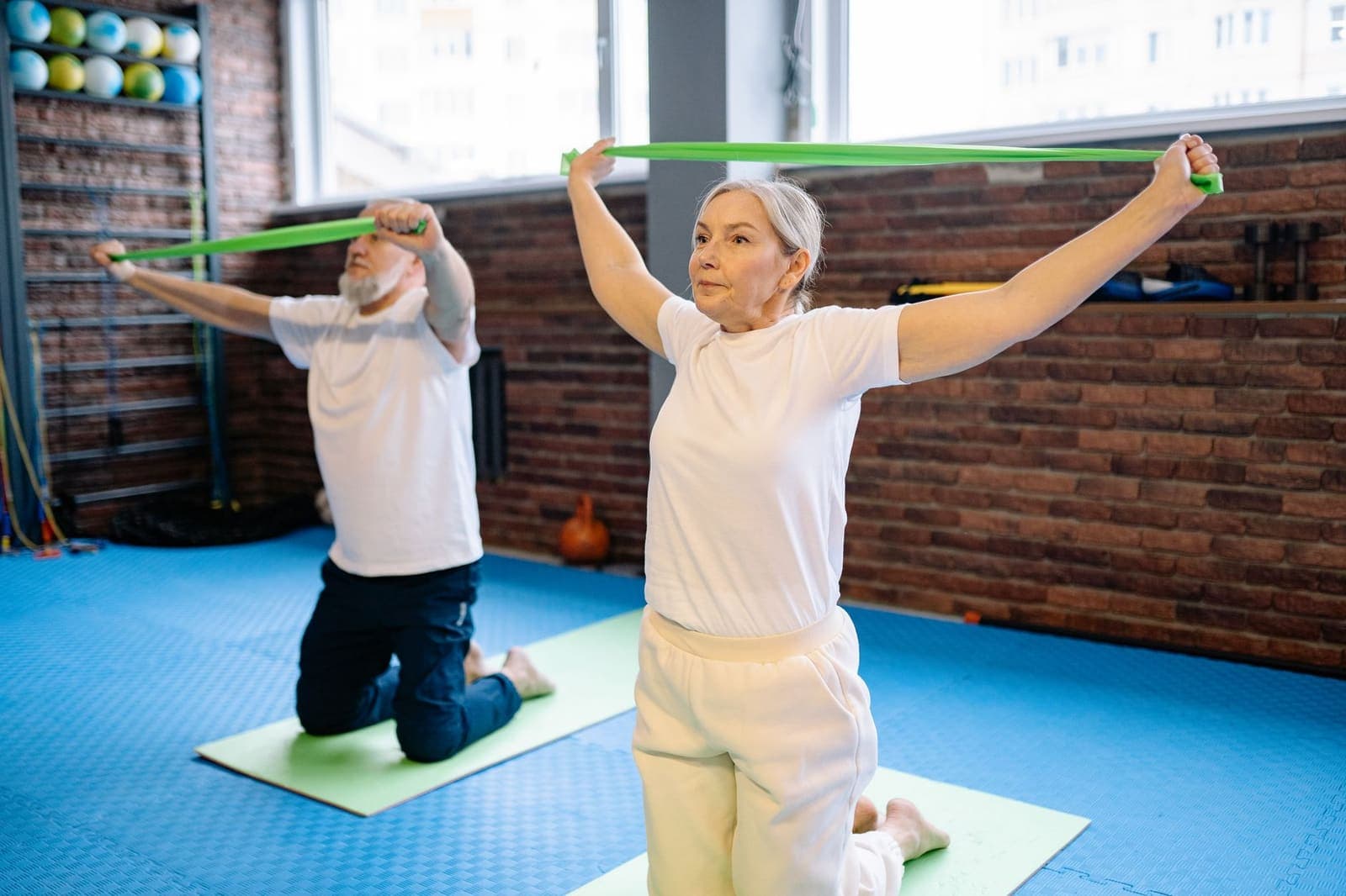 Two older adults kneel on exercise mats and stretch green resistance bands overhead in a brick-walled therapy gym