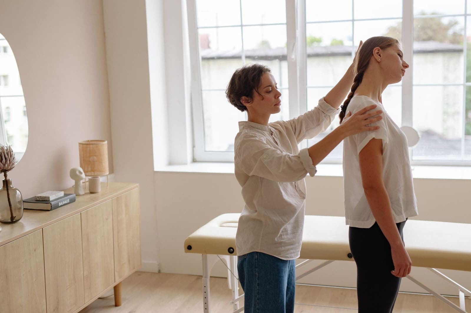 A practitioner places a hand on a standing patient's shoulder and adjusts her posture beside a treatment table