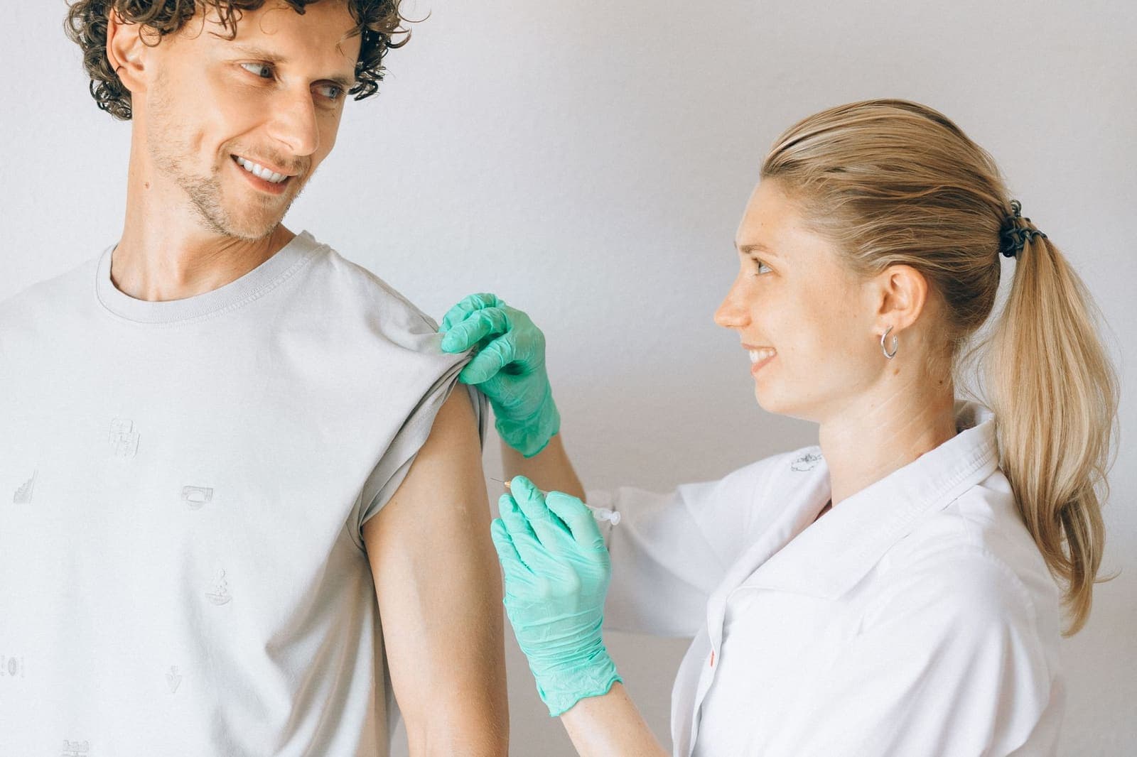 A clinician in a white coat and green gloves holds back a smiling patient's sleeve before giving a shot in the upper arm