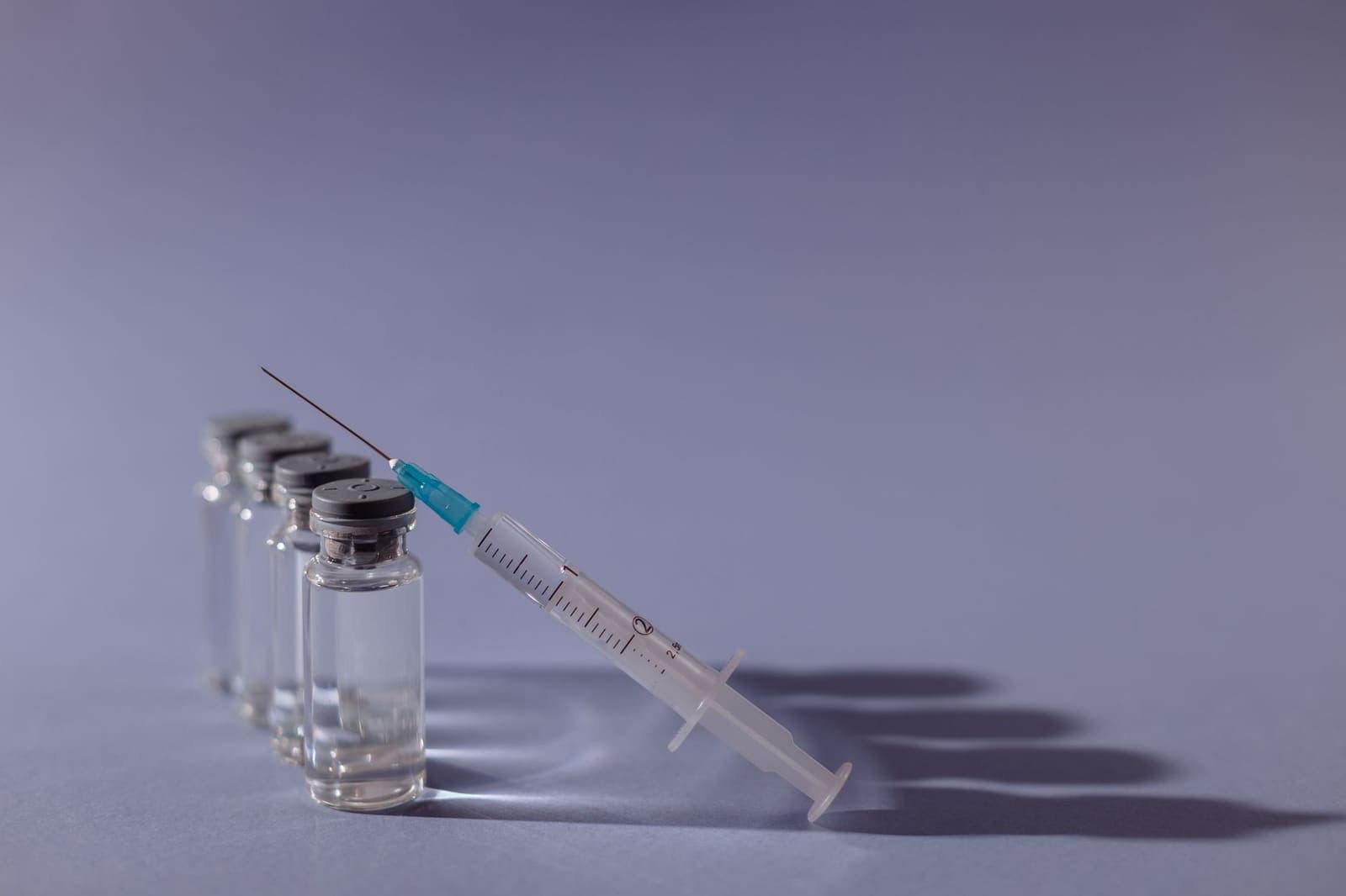 A row of four glass vaccine vials with a syringe leaning against the front one on a pale purple surface