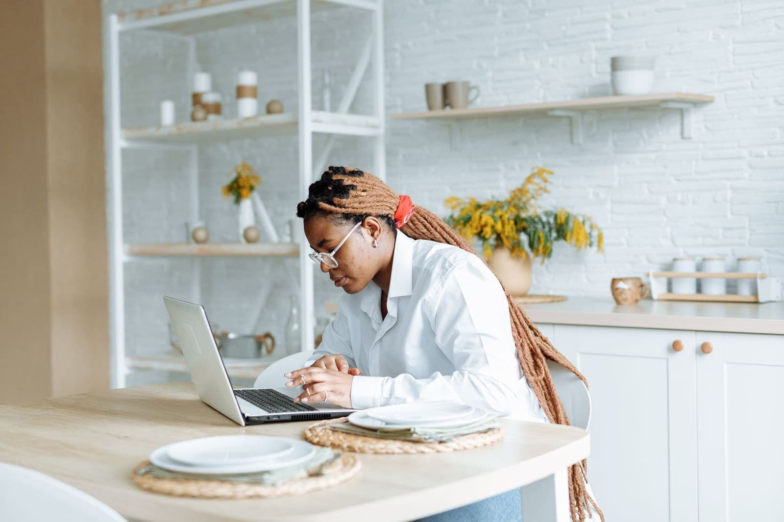 A woman in a white shirt types on a laptop at a kitchen table set with plates