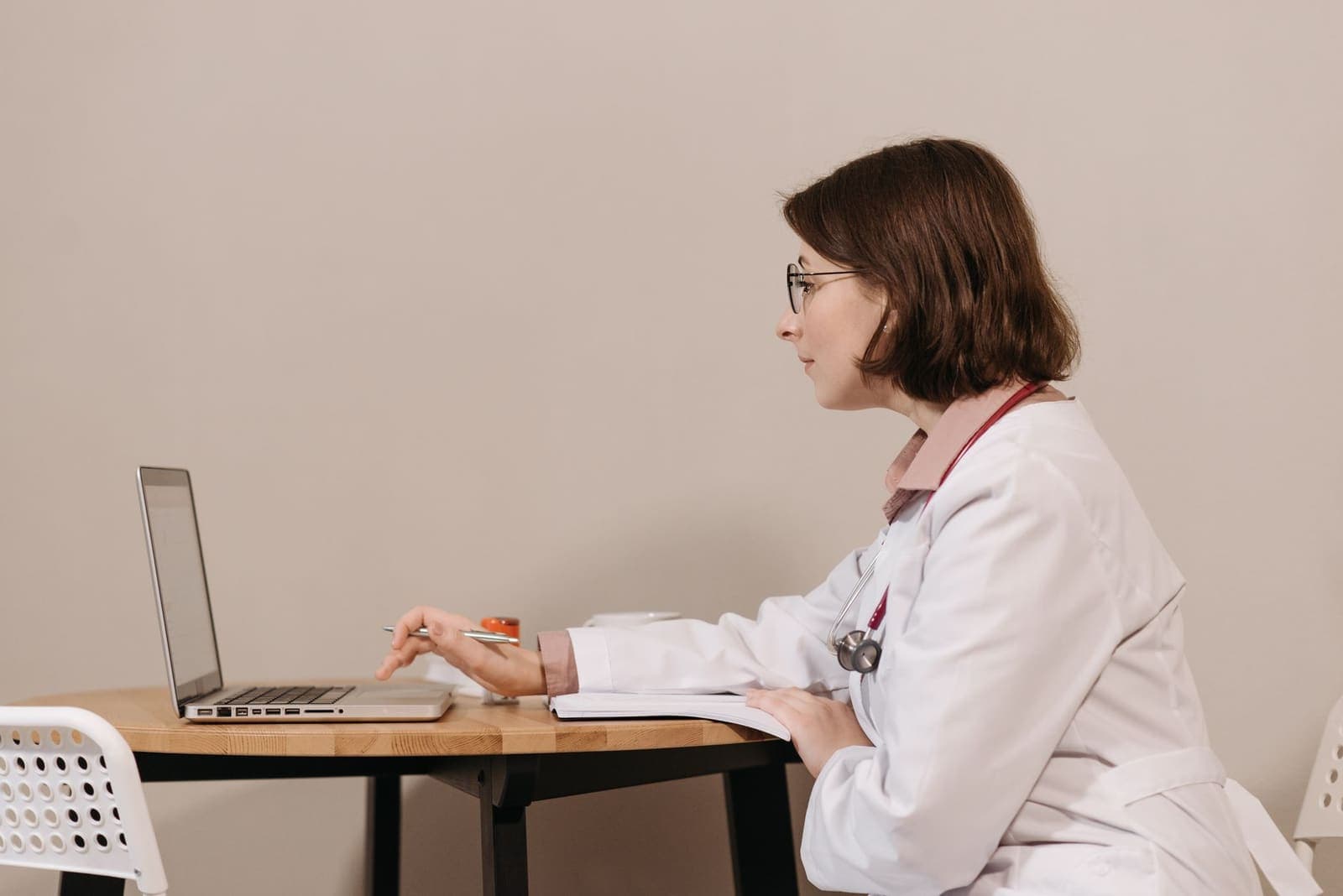A physician in a white coat with a stethoscope works at a laptop at a small table in a clinic