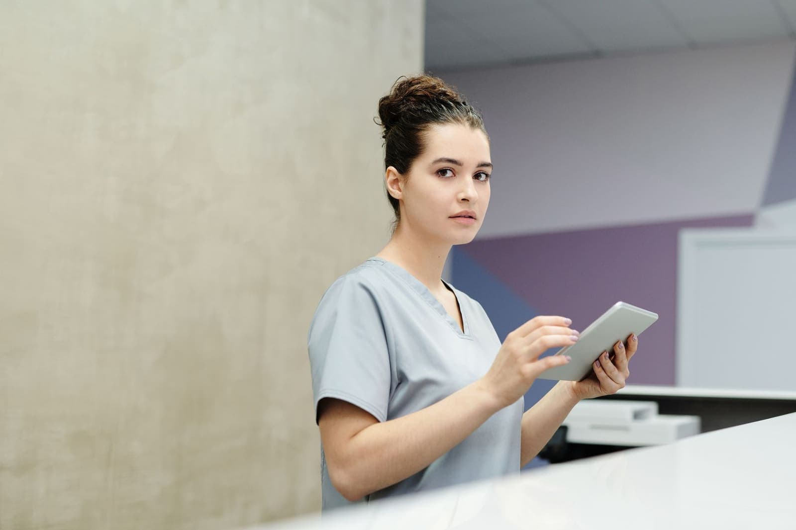 A receptionist in grey scrubs uses a tablet at a clinic front desk