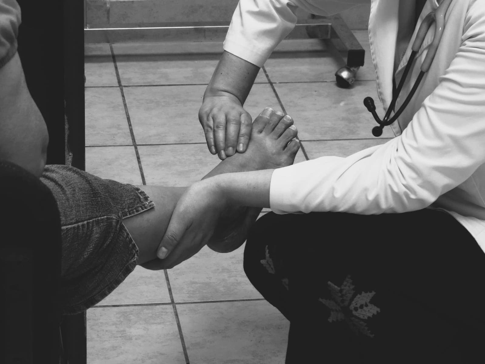 A black and white photograph of a clinician in a white coat examining a seated patient's bare foot