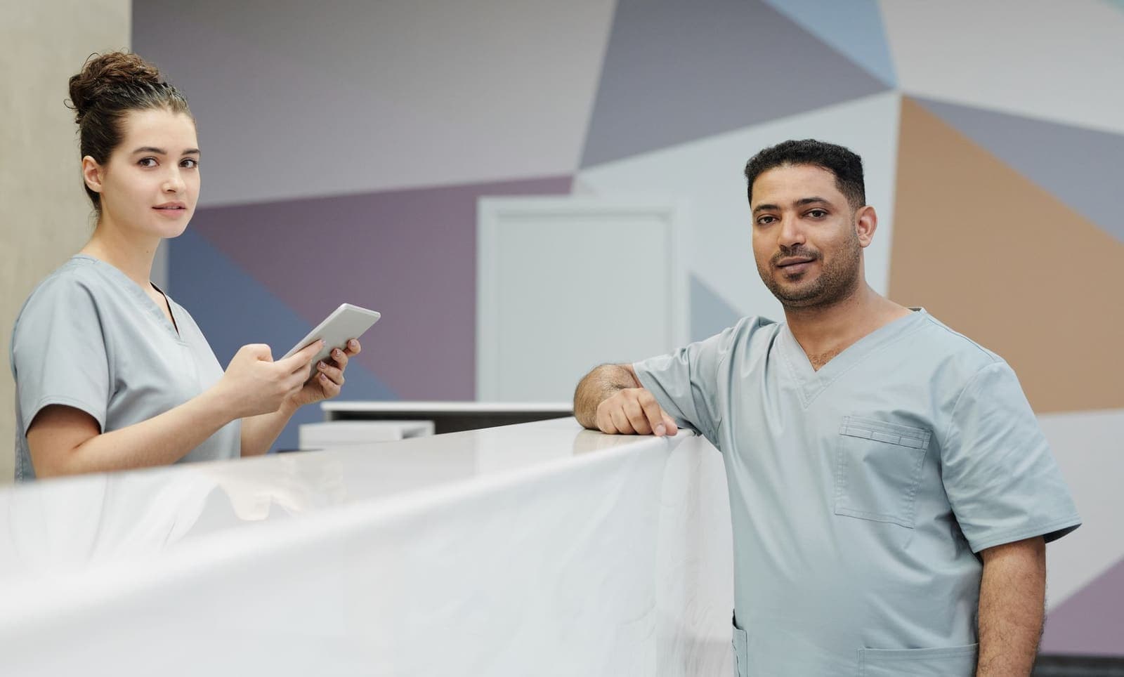Two staff members in grey scrubs stand at a white reception counter, one holding a tablet