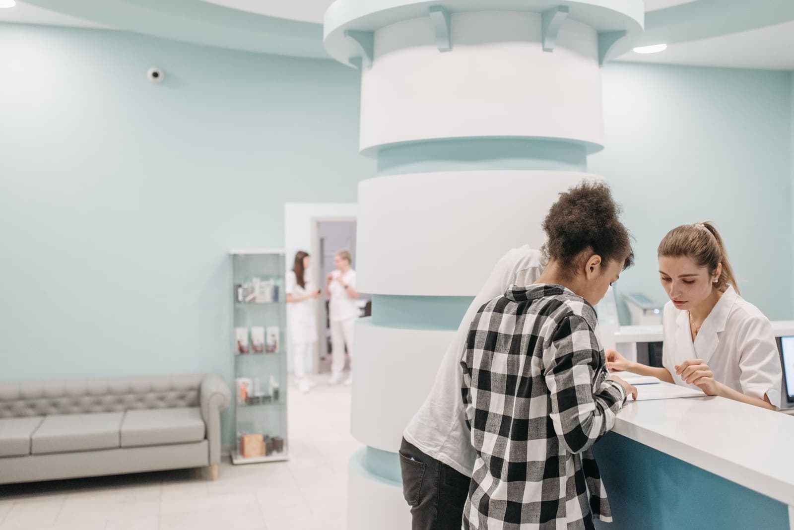 A patient standing at a clinic reception desk while a staff member in a white coat reviews a form with her