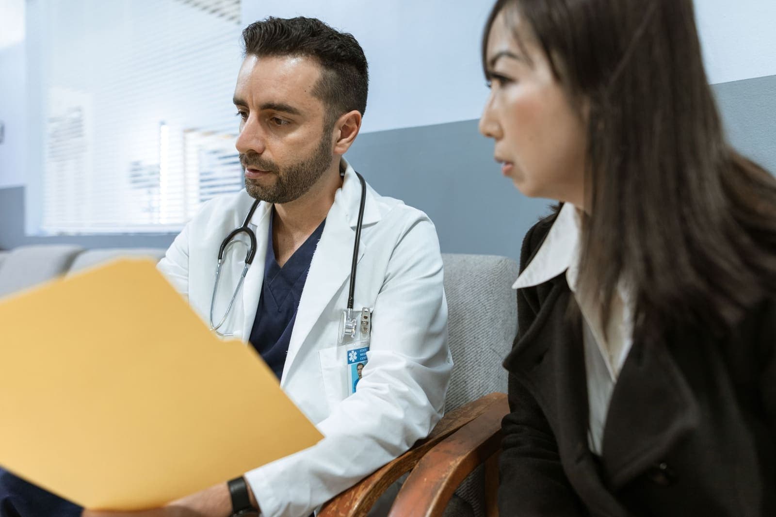 A doctor in a white coat and a woman in a dark coat sitting together, looking over papers in a yellow folder