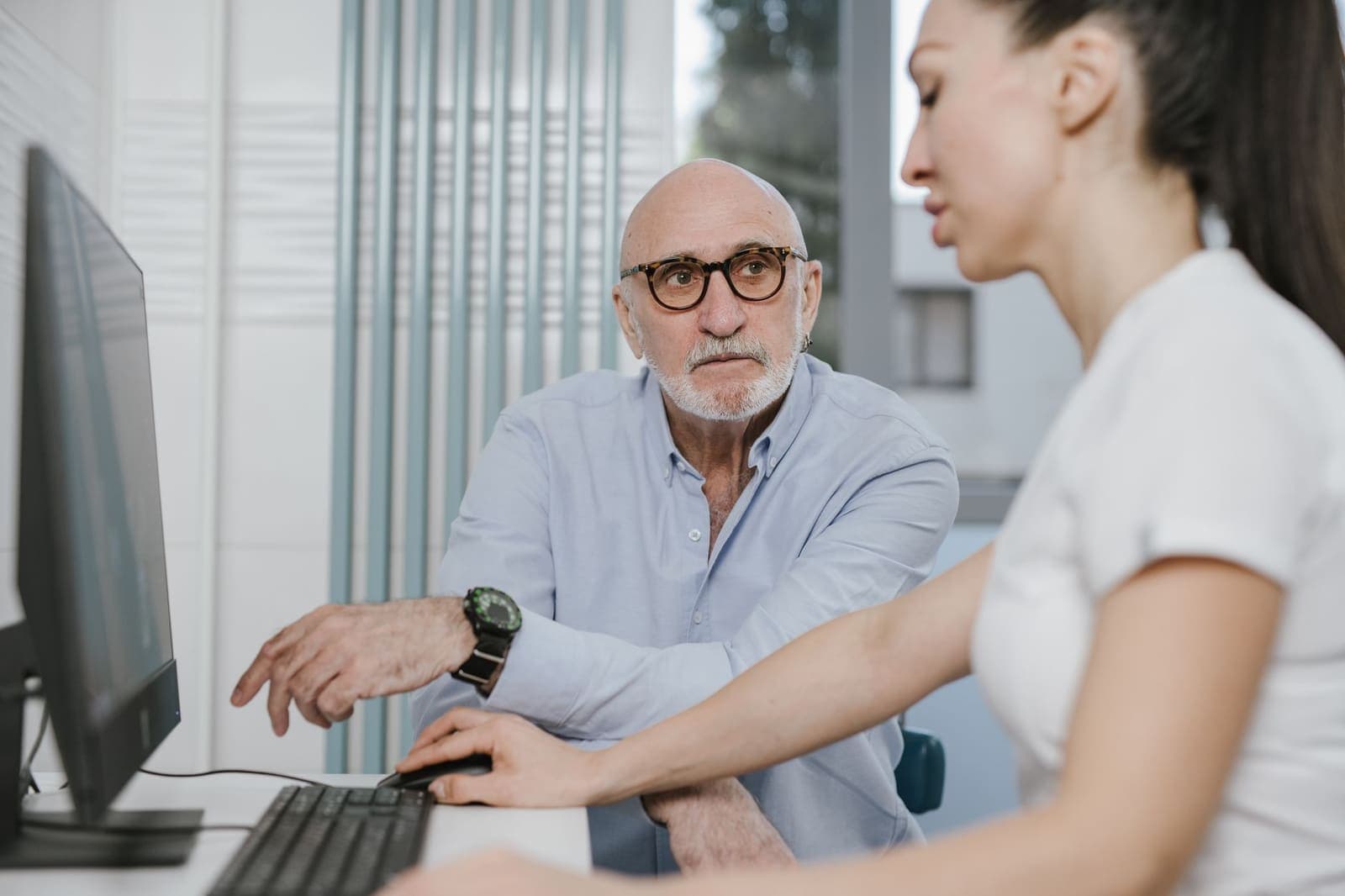 An older man in glasses at a practice desk pointing at a computer screen while a staff member works the mouse