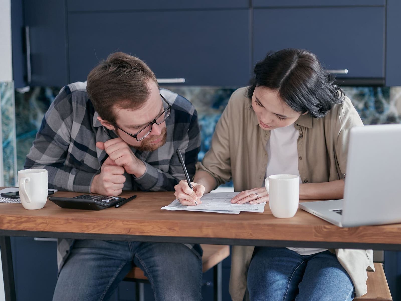A man and a woman at a kitchen table filling in a paper form beside a calculator and a laptop