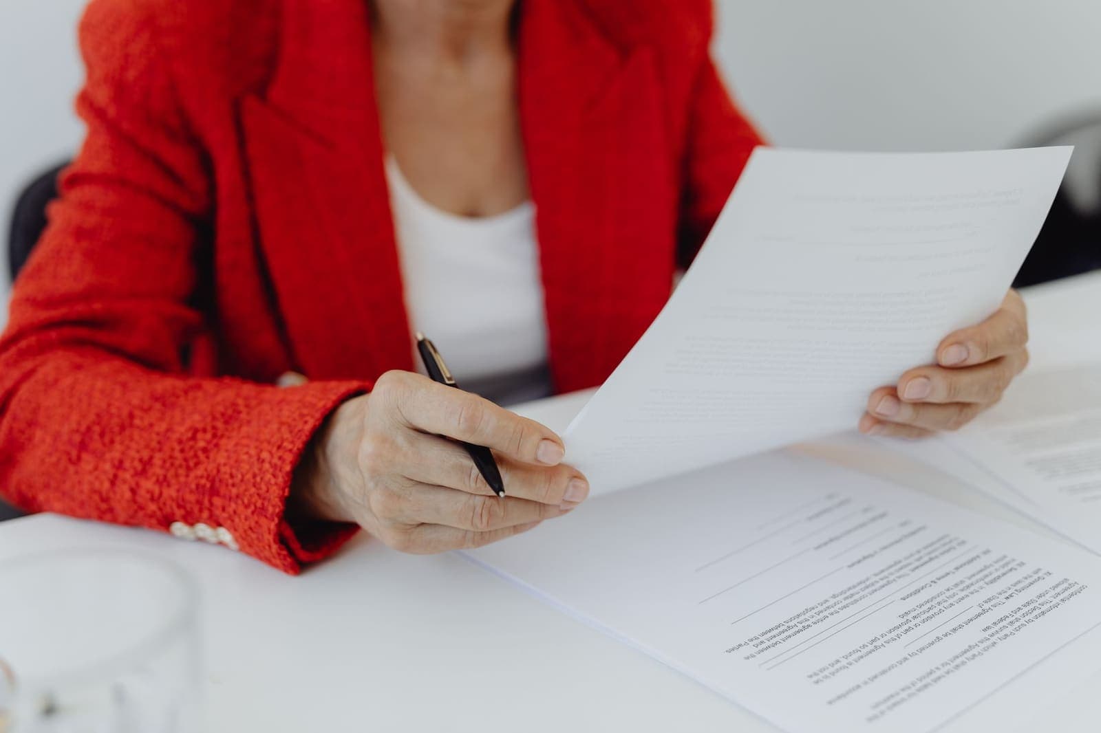 A woman in a red jacket holding a pen and reading a printed document at a white desk