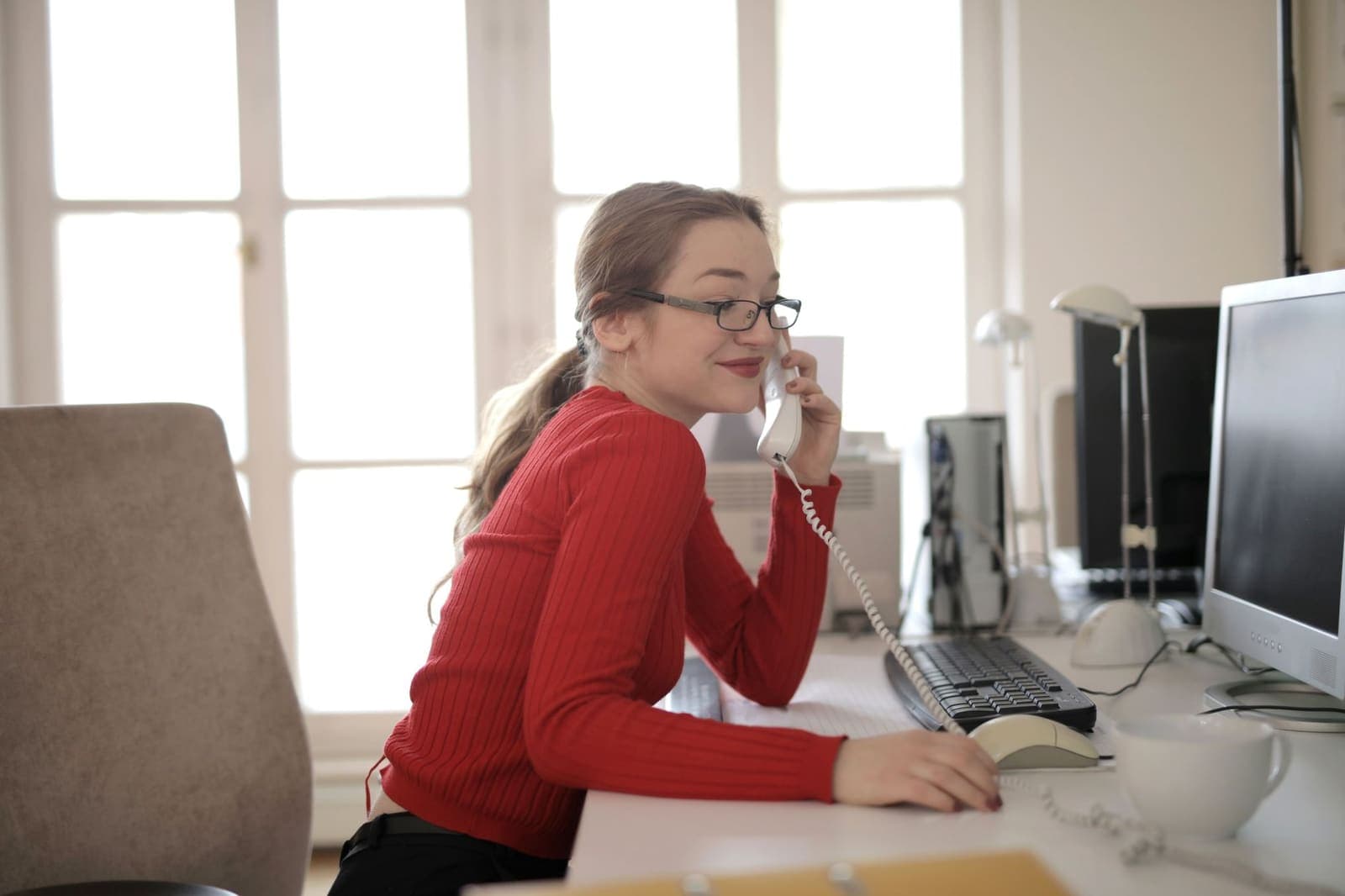 A woman in glasses and a red sweater talking on a desk phone in a bright office