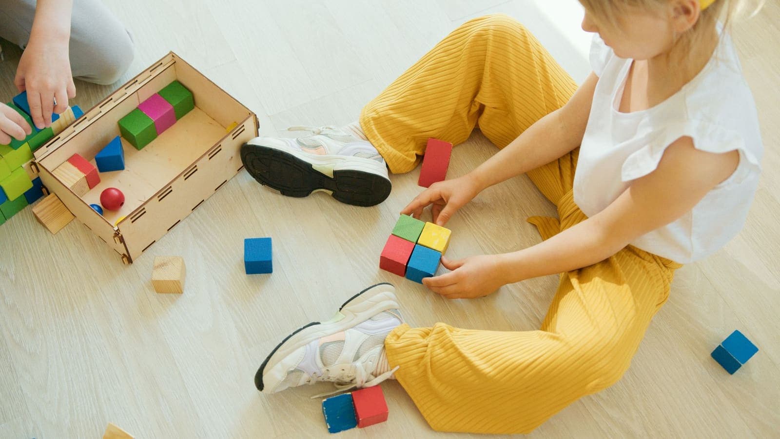 A young child in yellow trousers sitting on a light wooden floor, fitting four coloured blocks together beside a wooden box of blocks