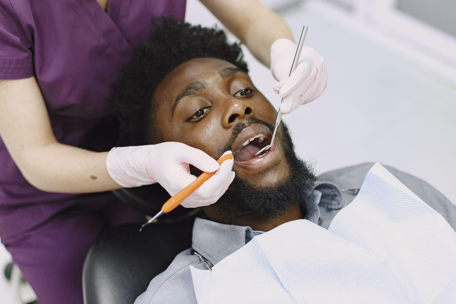 A man in a dental chair with his mouth open while a dentist in purple scrubs examines his teeth with a mirror and probe