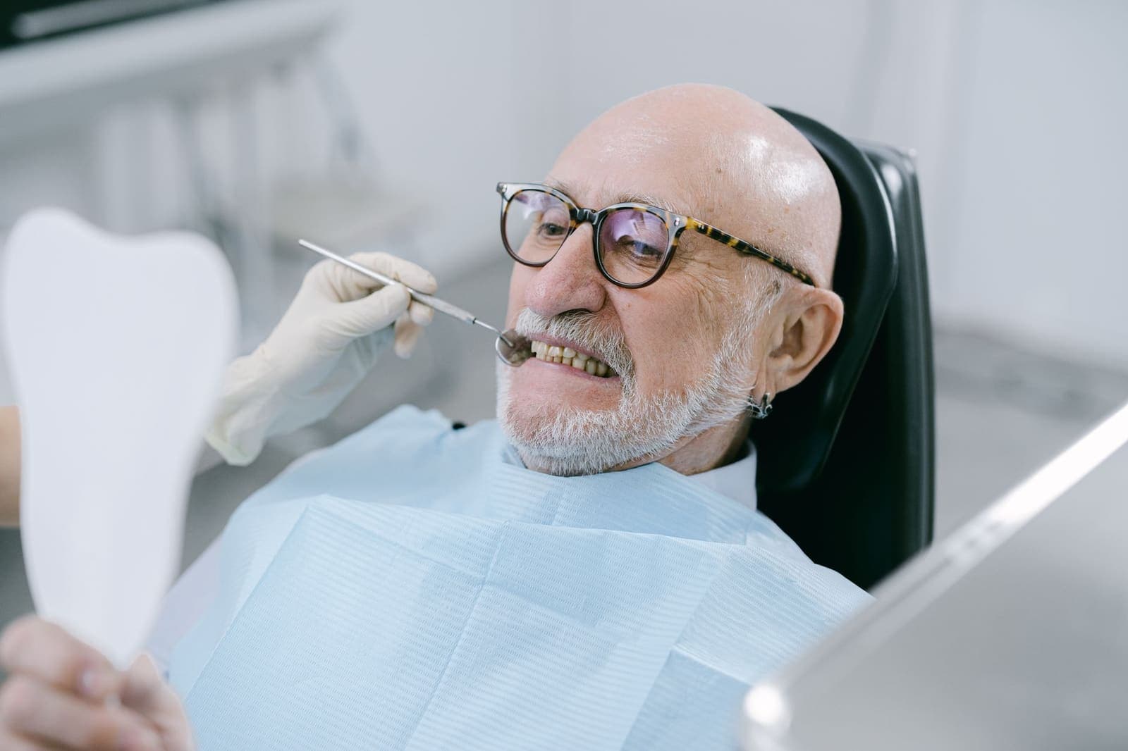 An older man in glasses reclining in a dental chair, looking at his teeth in a hand mirror while a gloved hand holds a dental mirror