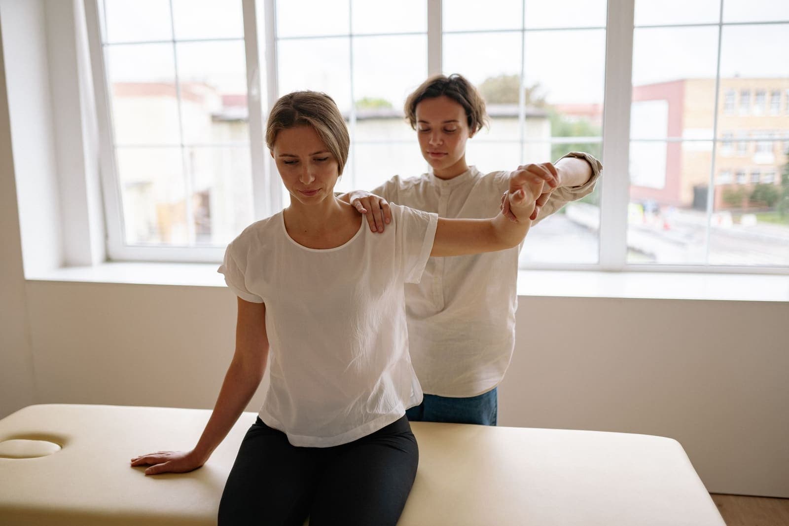 A therapist guiding a seated patient through a shoulder movement in a bright treatment room
