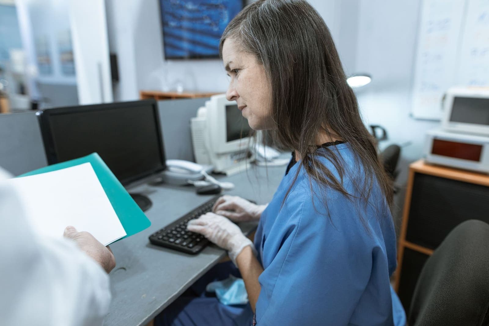 A clinical staff member typing at a desktop workstation while a colleague hands over a folder
