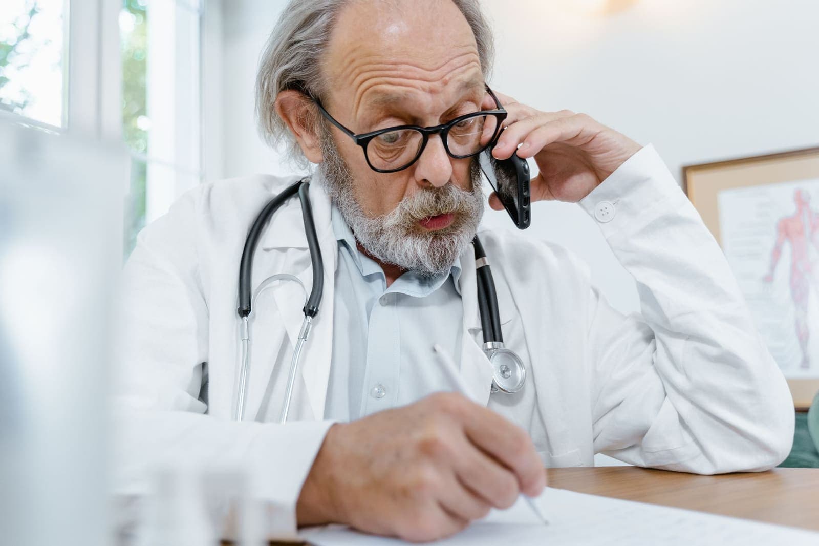 An older doctor with a grey beard and glasses talks on a mobile phone while writing on a form at his desk