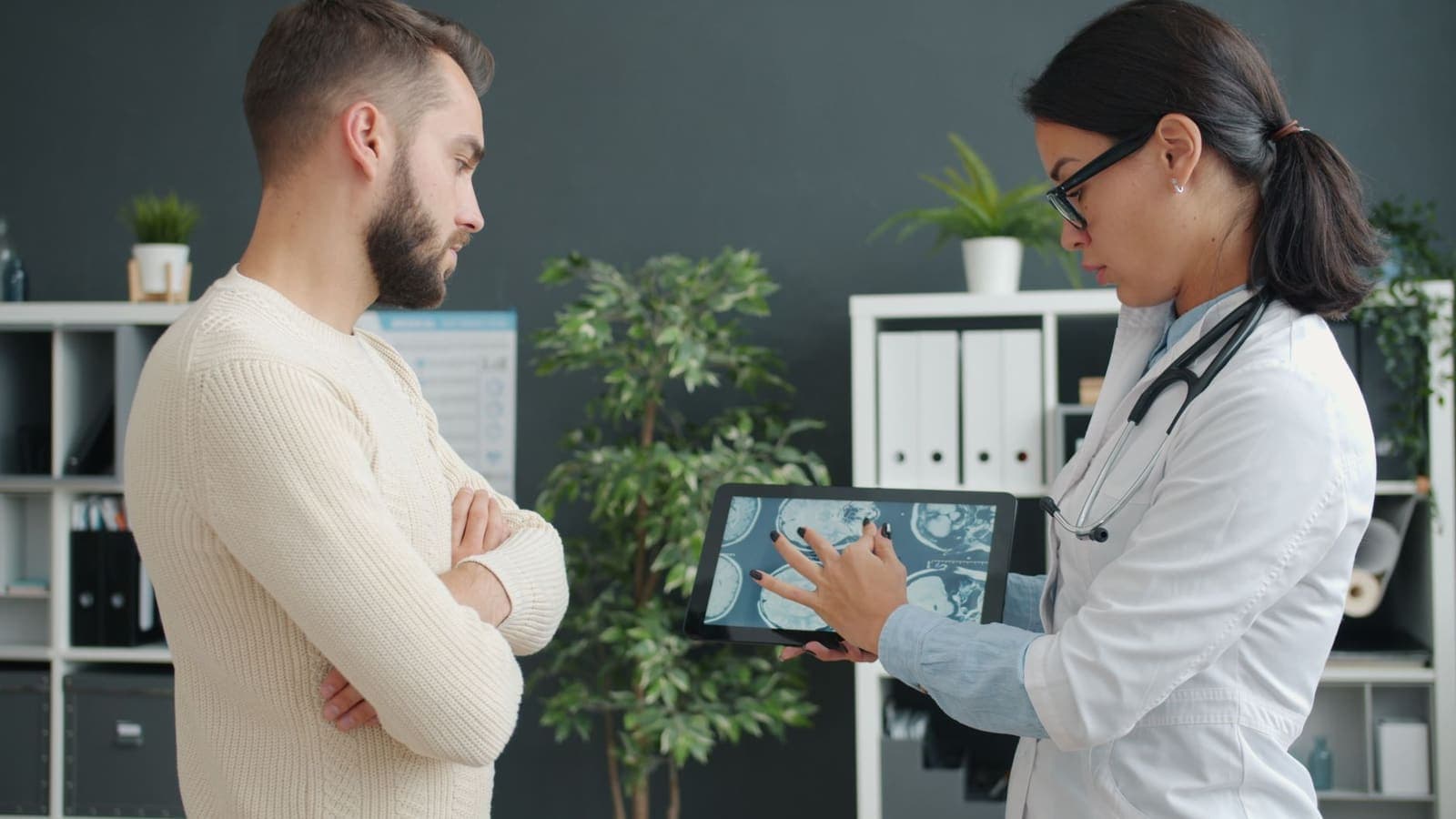 A doctor in a white coat shows MRI scan images on a tablet to a man in a cream sweater in a clinic office