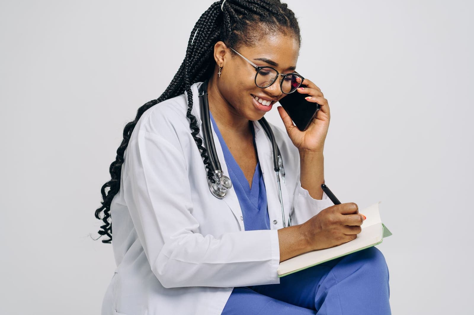 A doctor in a white coat and blue scrubs, a stethoscope round her neck, smiles while on a phone call and writes in a notebook