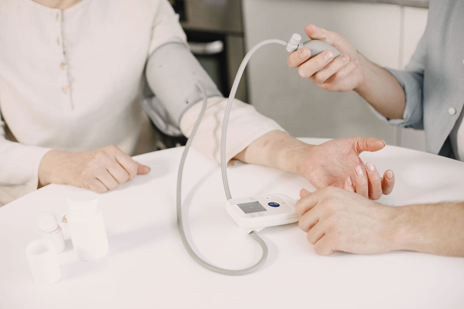 A clinician's hands hold a blood pressure cuff bulb while a patient's arm rests on a white table beside a digital monitor