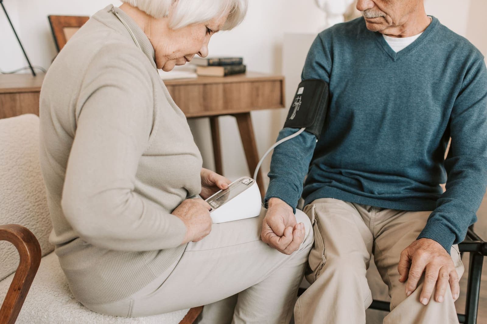 An older woman reads a home blood pressure monitor while the cuff is on her husband's arm in their living room