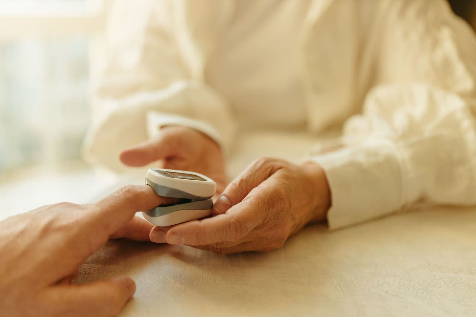 An older person's hands hold a pulse oximeter clipped onto another person's finger on a cream tablecloth