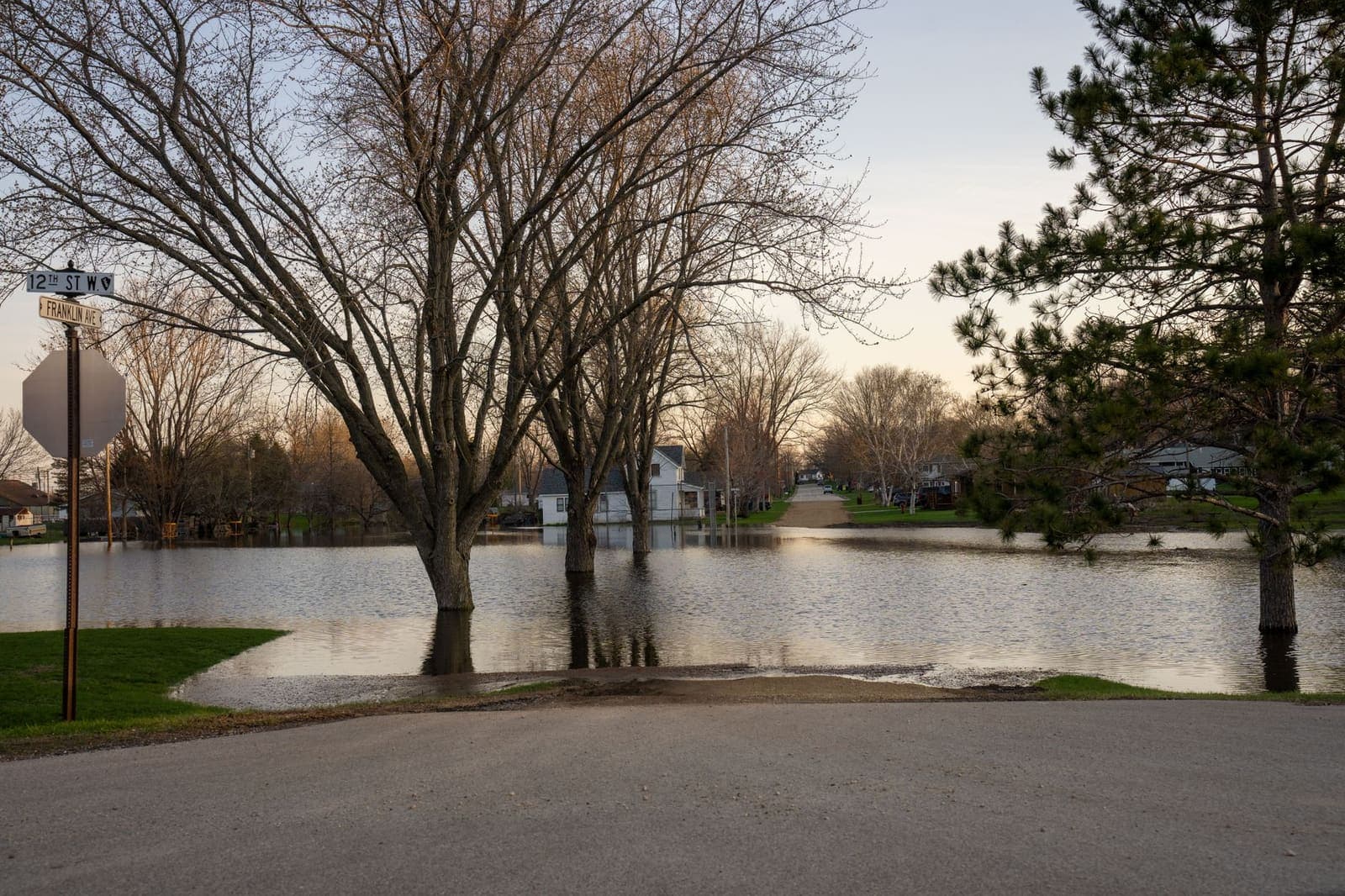 A residential street ending in standing floodwater with bare trees and houses behind it at dusk