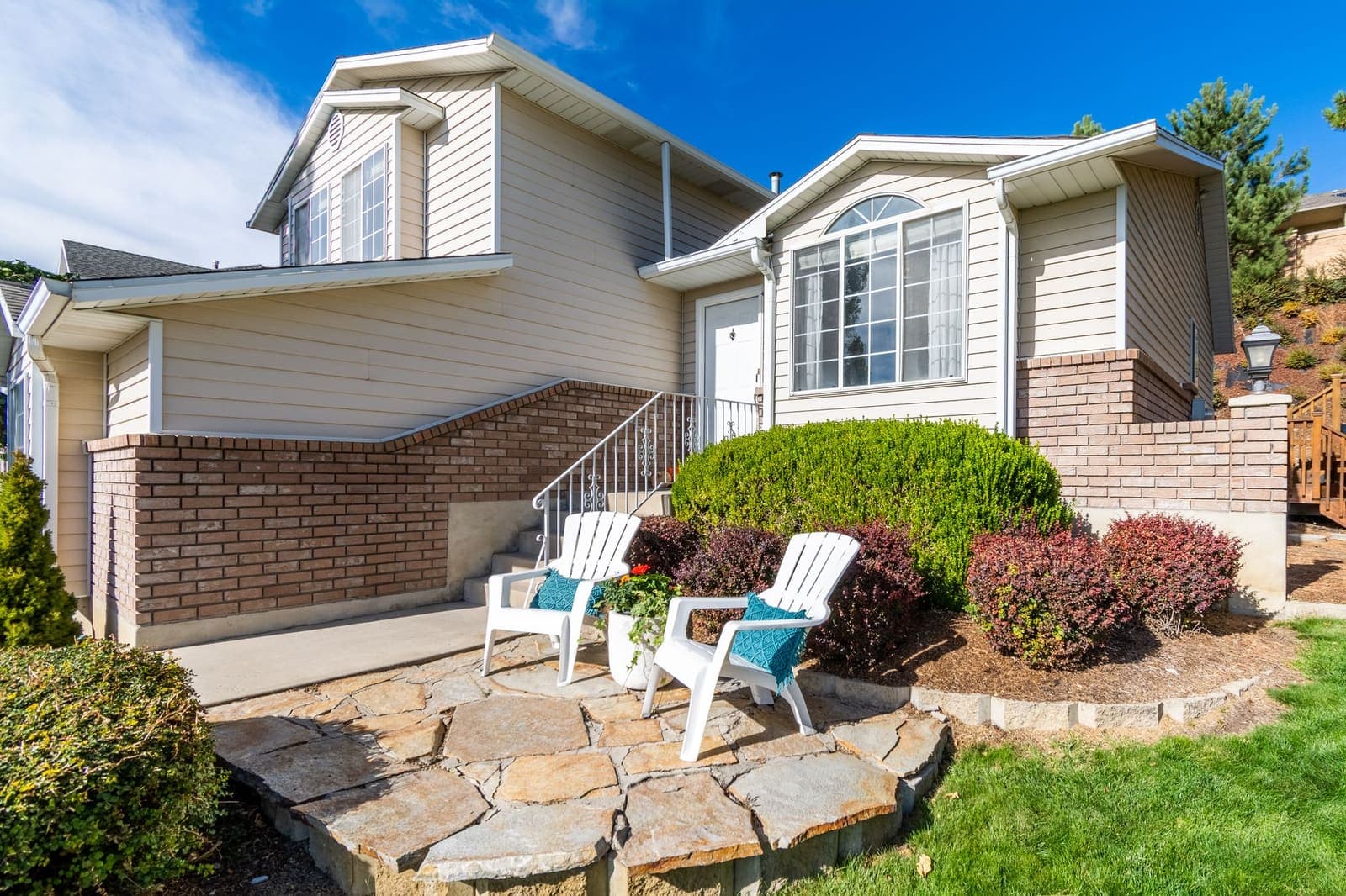 A split-level suburban house with a brick front, a small patio and two white chairs on a sunny day